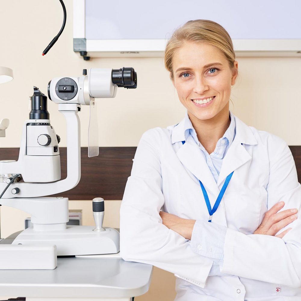 Portrait of blonde female ophthalmologist sitting at slit lamp machine while posing confidently with arms crossed and smiling happily